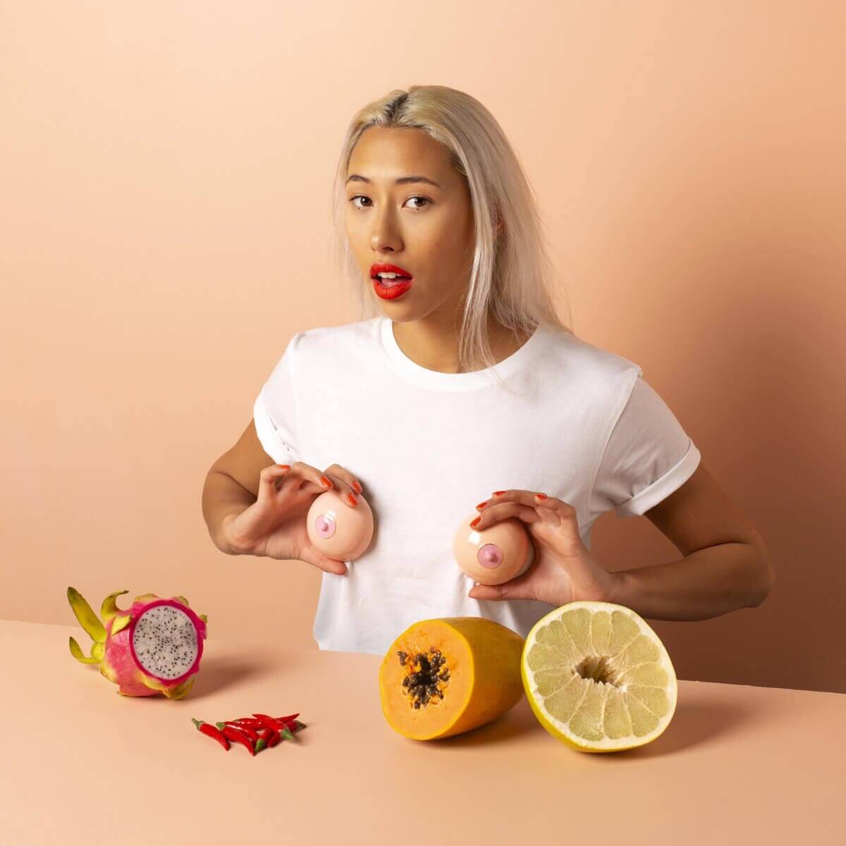 Woman wearing a white T-shirt aand standing in front of a table covered with exotic fruits, holding two breast-shaped enamel boxes over her breast area Nudie Co