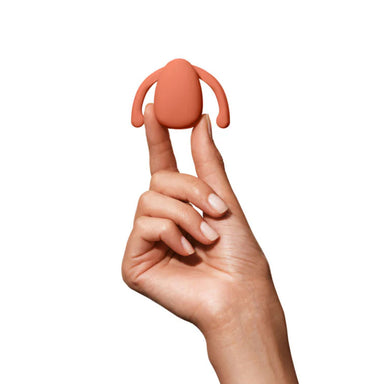 Hand holding an orange silicone vibrator for clitoris with two wings on the side to nest inside labia Nudie Co