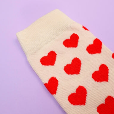 Close -up of a cream cotton sock with red heart print over a purple background Nudie Co