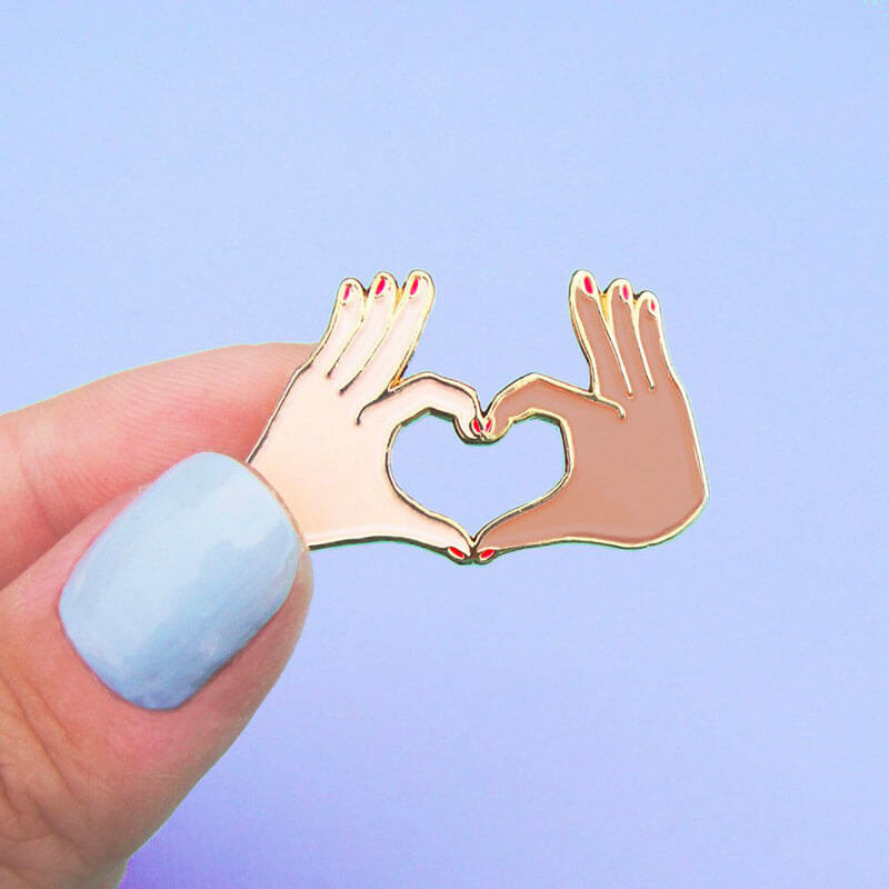 Fingers holding an enamel pin with two hands forming a love heart Nudie Co