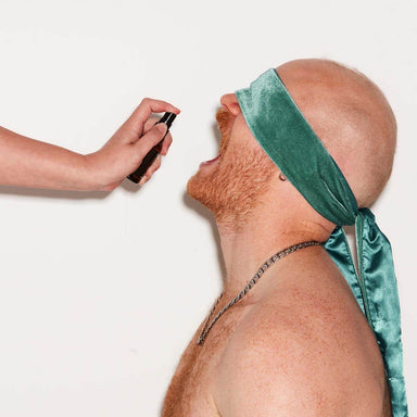 hand holding a black spray bottle over a person wearing green velvet blindfolds with an open mouth Nudie Co