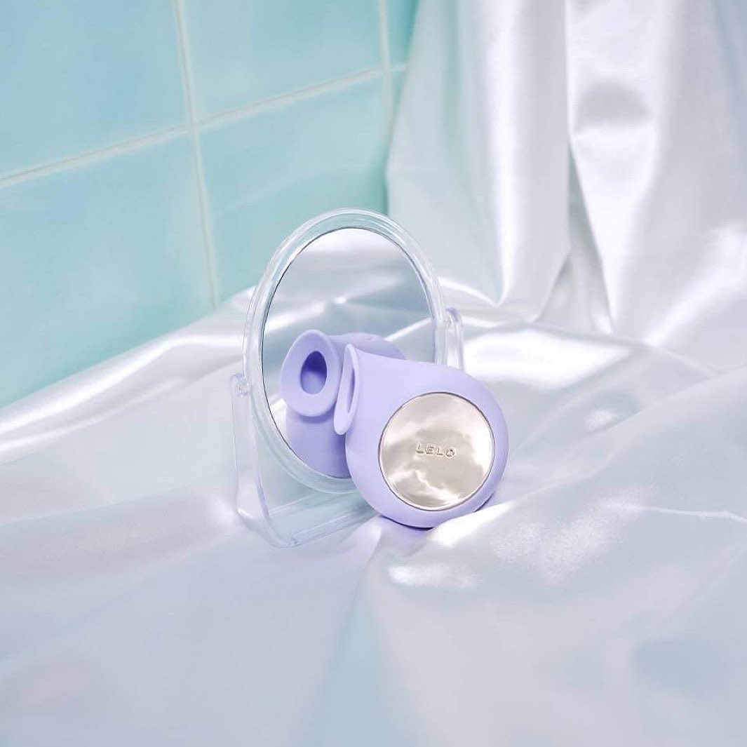 Purple round Lelo Sila clitoral masssger sitting on a white satin fabric and resting against a small mirror in a blue-tiled bathroom Nudie Co