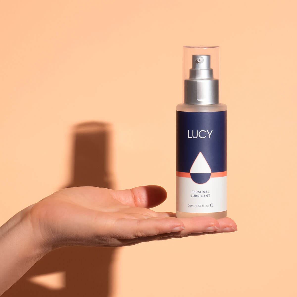Woman's hand holding a glass pump bottle of water-based lubricant over a peach-coloured wall Nudie Co