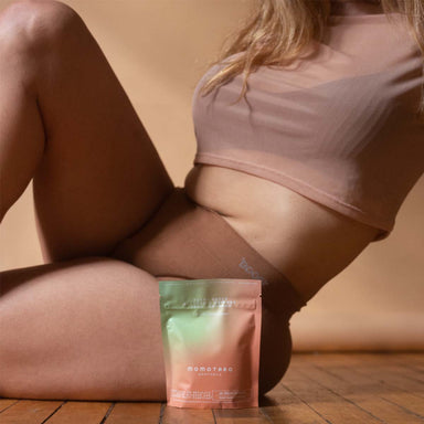 Woman in underwear sitting on a wooden floor with a Momotaro Apotheca packet of UTI supplement Nudie Co