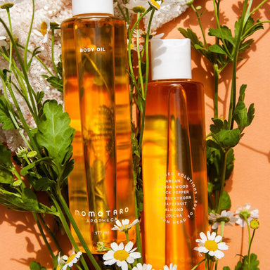 Two bottles of Momotaro Apotheca body oil over a peach-coloured background with wild flowers Nudie Co