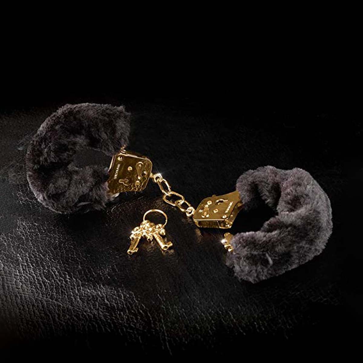 Black fluffy handcuffs with gold metal details and two gold metal keys over a black textured background Nudie Co