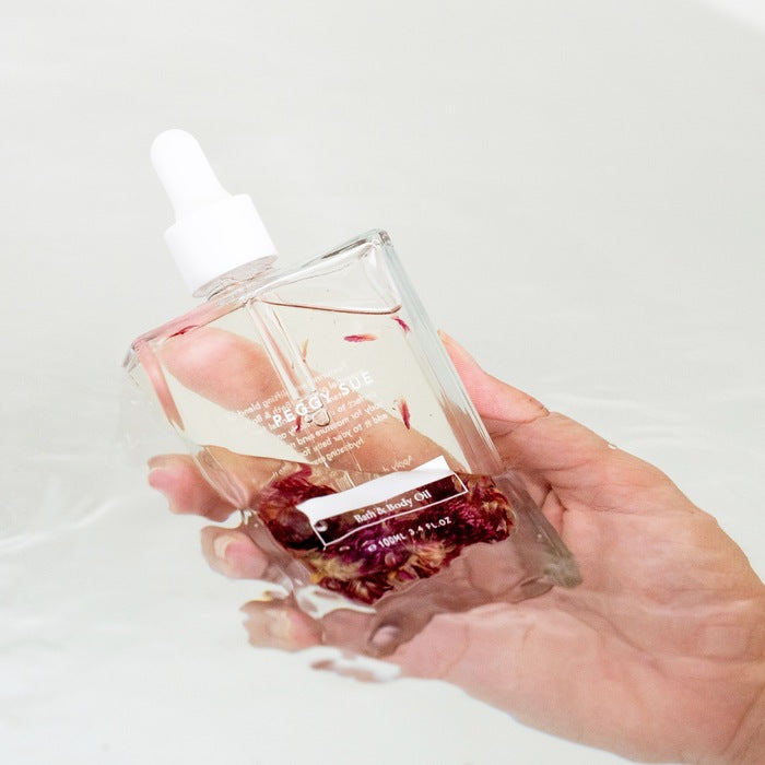 Woman holding a bottle of vegan and organic bath and body oil wth dried flowers in a bath Nudie Co