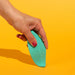 Woman's hand pushing the tip of a green leaf-shaped silicone vibrator against a bright yellow surface Nudie Co